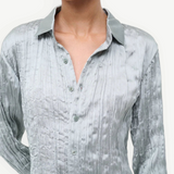 A person wearing the Suki Shirt, a sage green button-up with a long-sleeved, crinkled texture that serves as a versatile wardrobe essential.