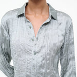 A person wearing the Suki Shirt, a sage green button-up with a long-sleeved, crinkled texture that serves as a versatile wardrobe essential.