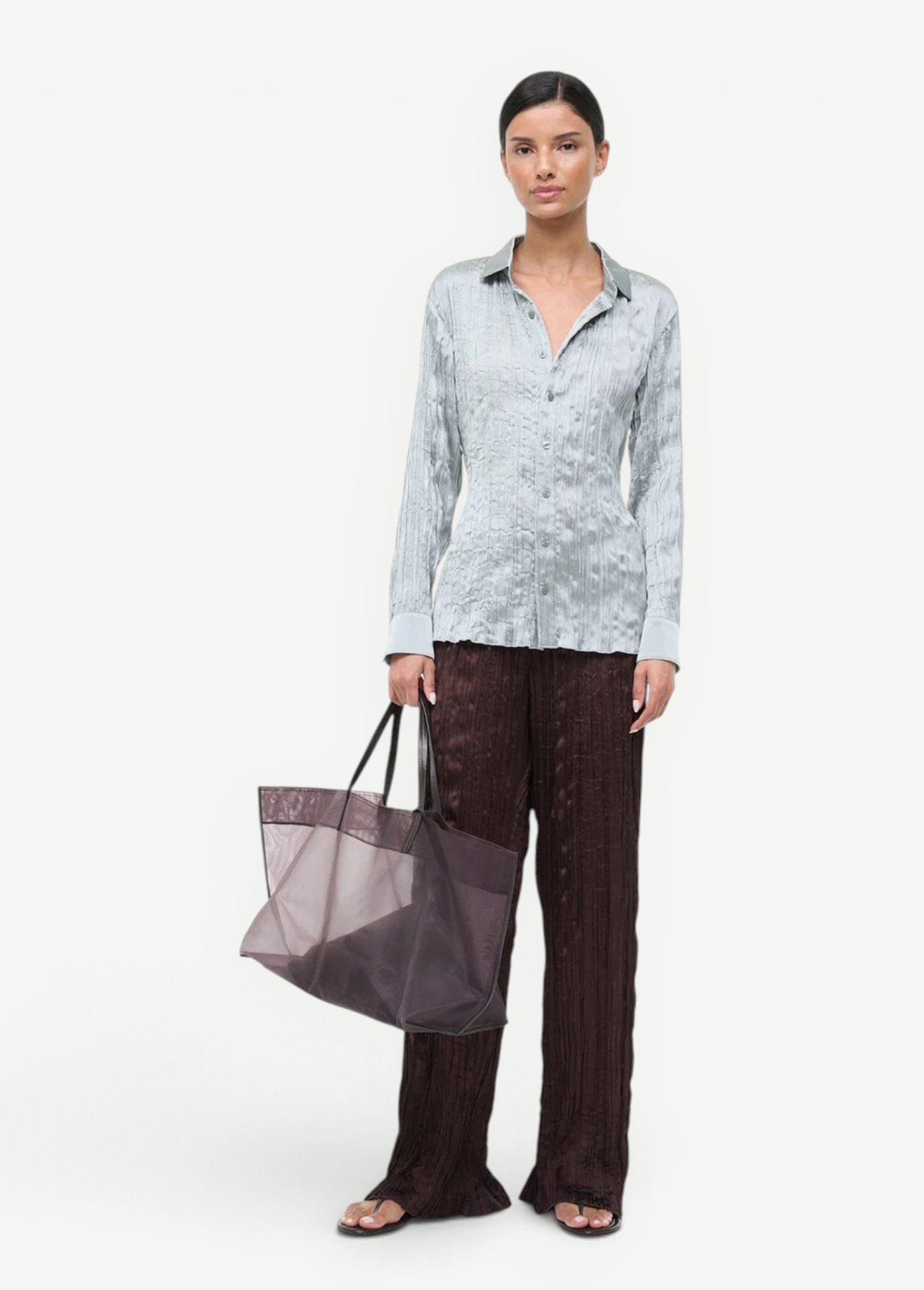 A person wears a light blue, textured Suki Shirt and dark brown textured trousers while holding a large translucent tote bag, an effortless styling choice.