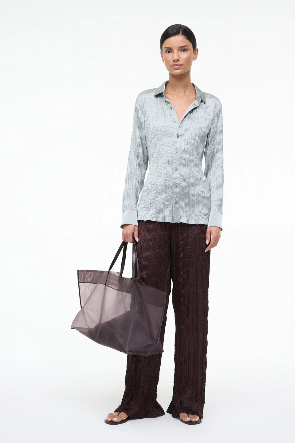 A person wears a light blue, textured Suki Shirt and dark brown textured trousers while holding a large translucent tote bag, an effortless styling choice.