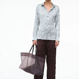 A person wears a light blue, textured Suki Shirt and dark brown textured trousers while holding a large translucent tote bag, an effortless styling choice.