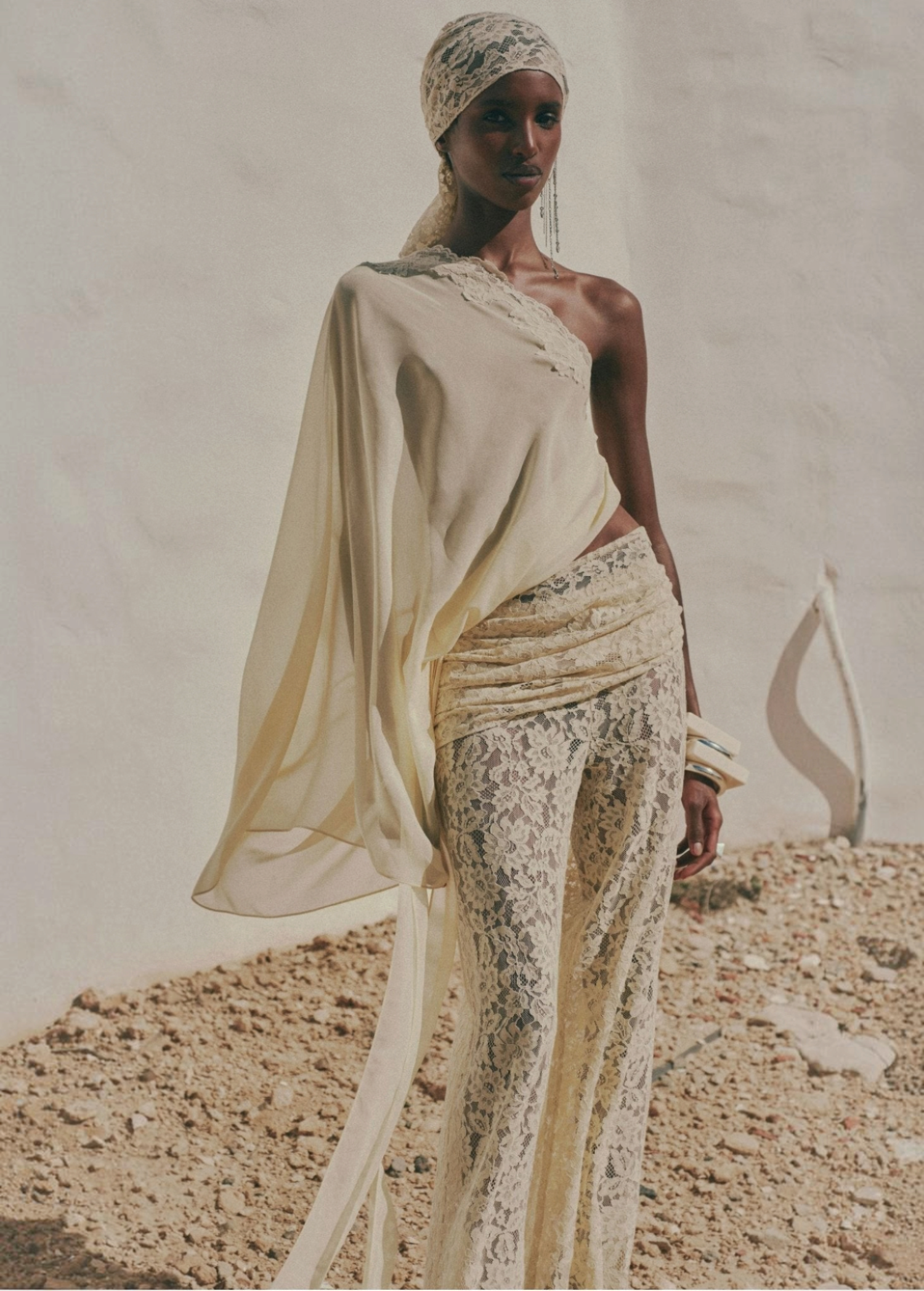 A person wears a one-shoulder pale yellow top with a draped sleeve, paired with vintage-inspired Rocio Trouser lace trousers and a matching lace headpiece.