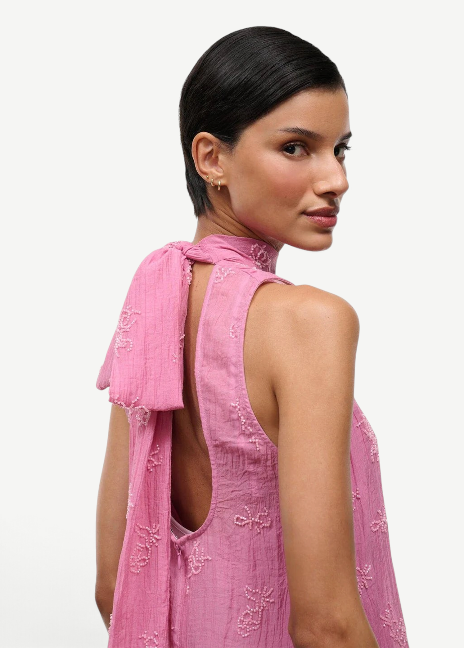 A person wears the pink Mini Albee Dress, an elegant mini dress featuring a sophisticated silhouette with a high neck, open back, and beaded floral embroidery.