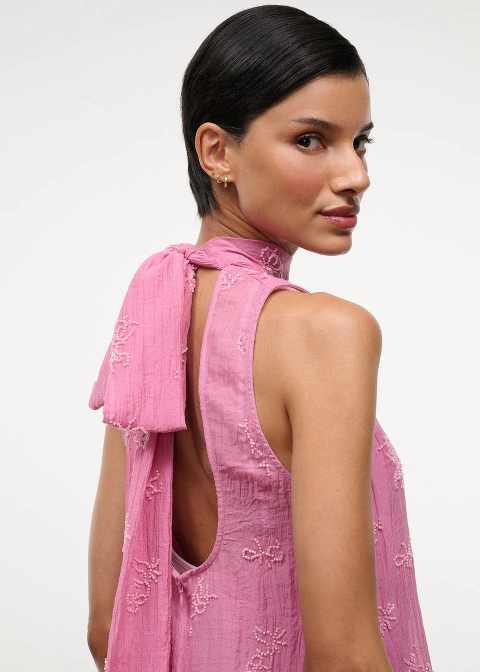 A person wears the pink Mini Albee Dress, an elegant mini dress featuring a sophisticated silhouette with a high neck, open back, and beaded floral embroidery.