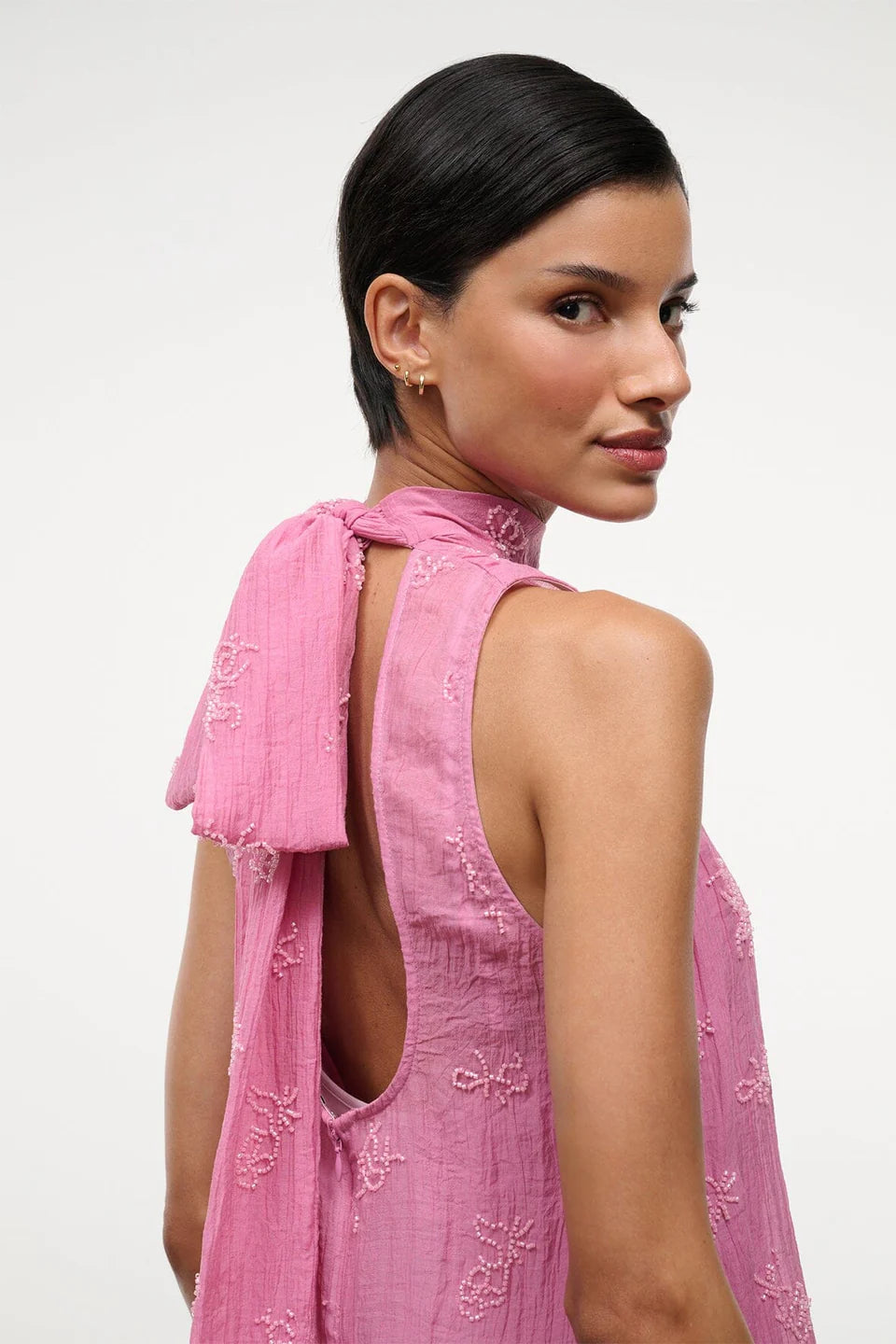 A person wears the pink Mini Albee Dress, an elegant mini dress featuring a sophisticated silhouette with a high neck, open back, and beaded floral embroidery.
