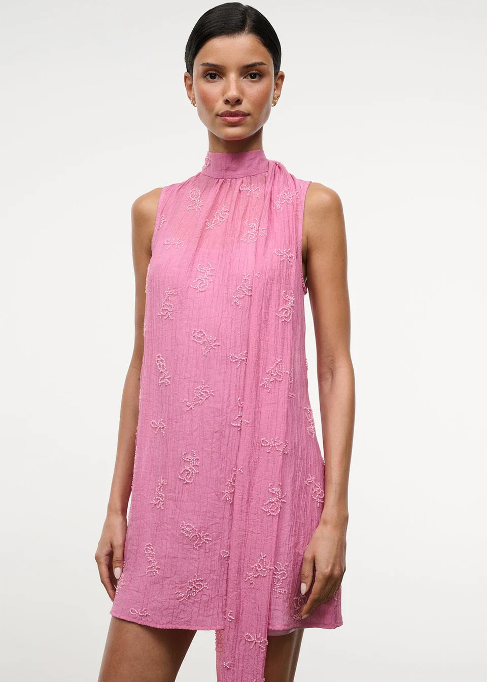 The Mini Albee Dress is a sleeveless, high-neck mini dress in pink featuring textured floral embroidery and an attached side scarf for a sophisticated silhouette.