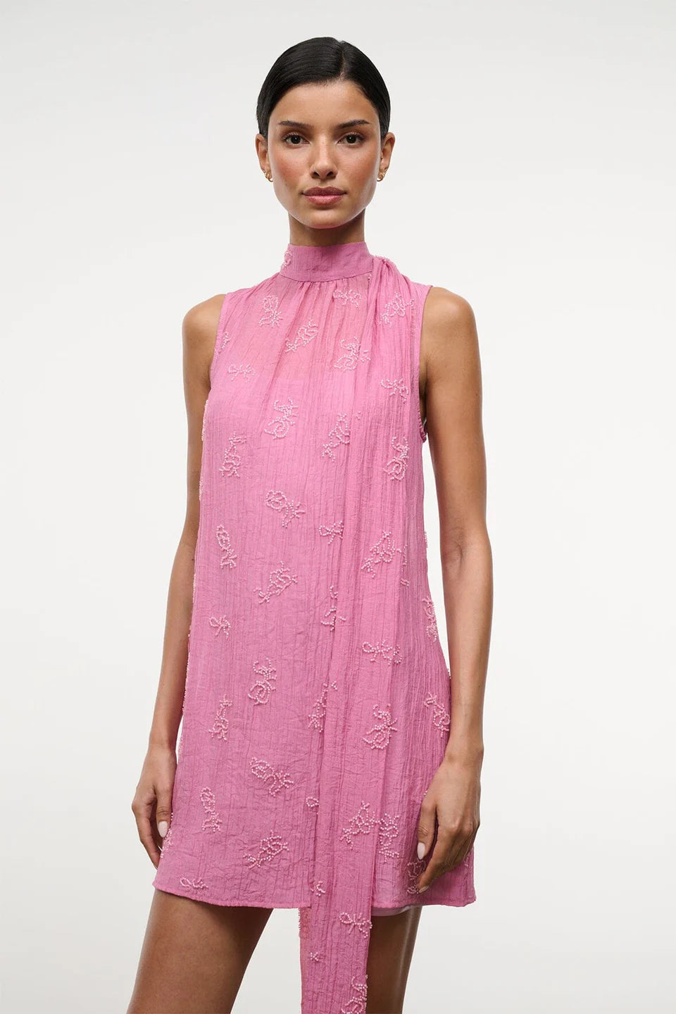 The Mini Albee Dress is a sleeveless, high-neck mini dress in pink featuring textured floral embroidery and an attached side scarf for a sophisticated silhouette.