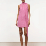 A model wears the Mini Albee Dress, an elegant pink sleeveless mini dress with a high mock neck and a draped fabric panel extending from the shoulder, creating a sophisticated silhouette.