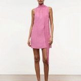 A model wears the Mini Albee Dress, an elegant pink sleeveless mini dress with a high mock neck and a draped fabric panel extending from the shoulder, creating a sophisticated silhouette.