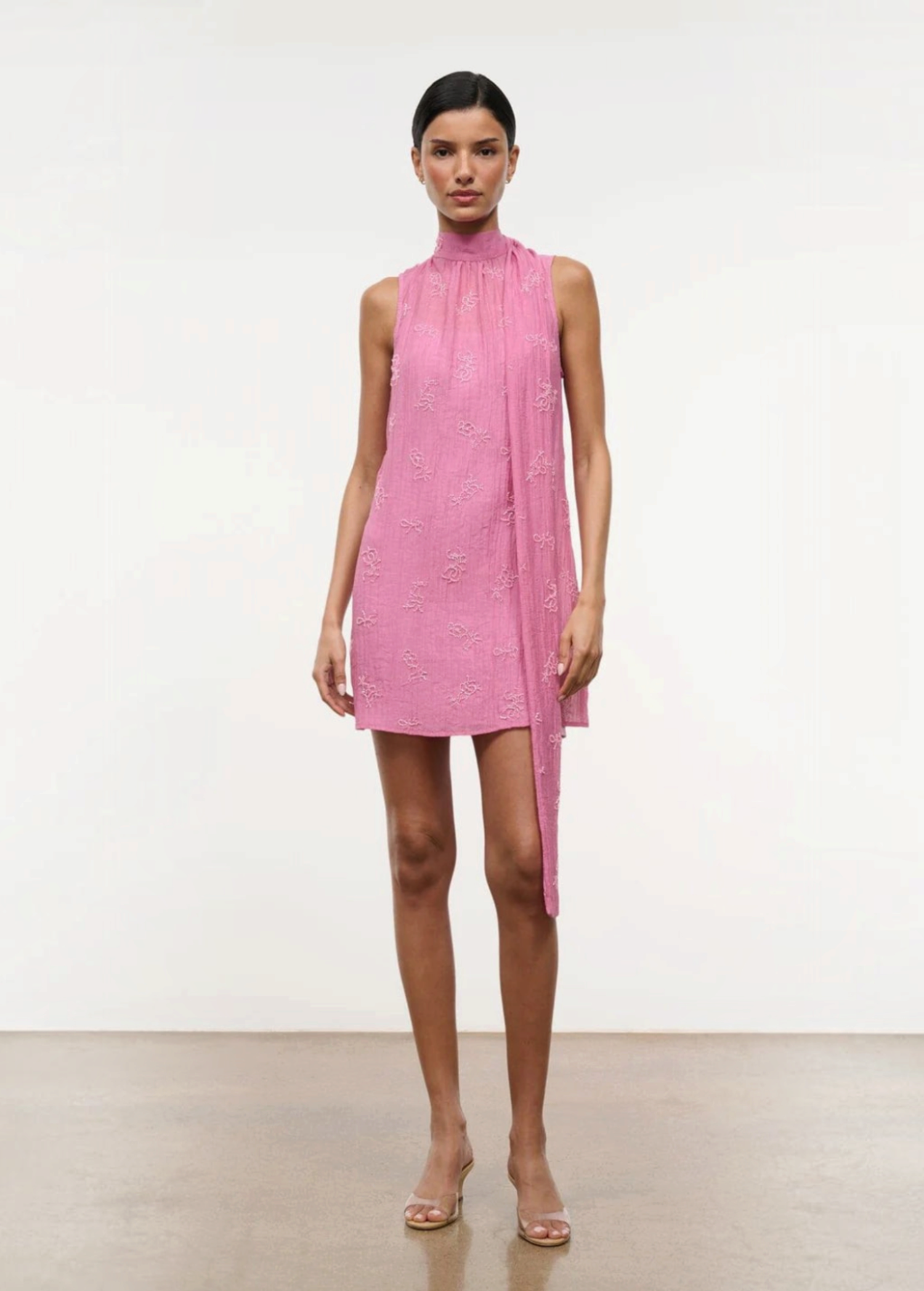 A model wears the Mini Albee Dress, an elegant pink sleeveless mini dress with a high mock neck and a draped fabric panel extending from the shoulder, creating a sophisticated silhouette.