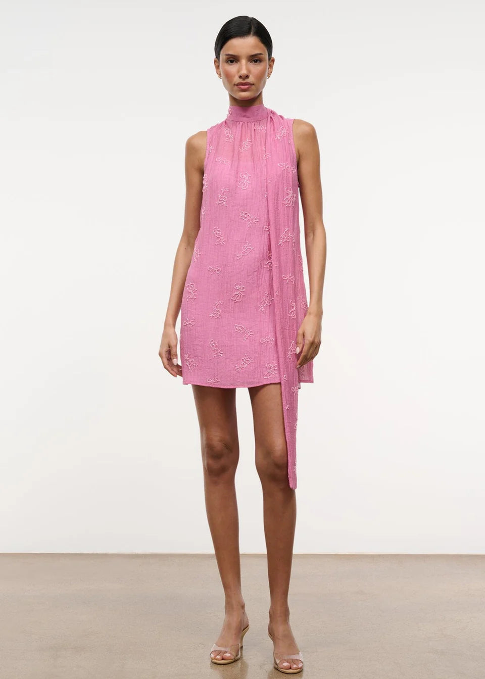 A model wears the Mini Albee Dress, an elegant pink sleeveless mini dress with a high mock neck and a draped fabric panel extending from the shoulder, creating a sophisticated silhouette.