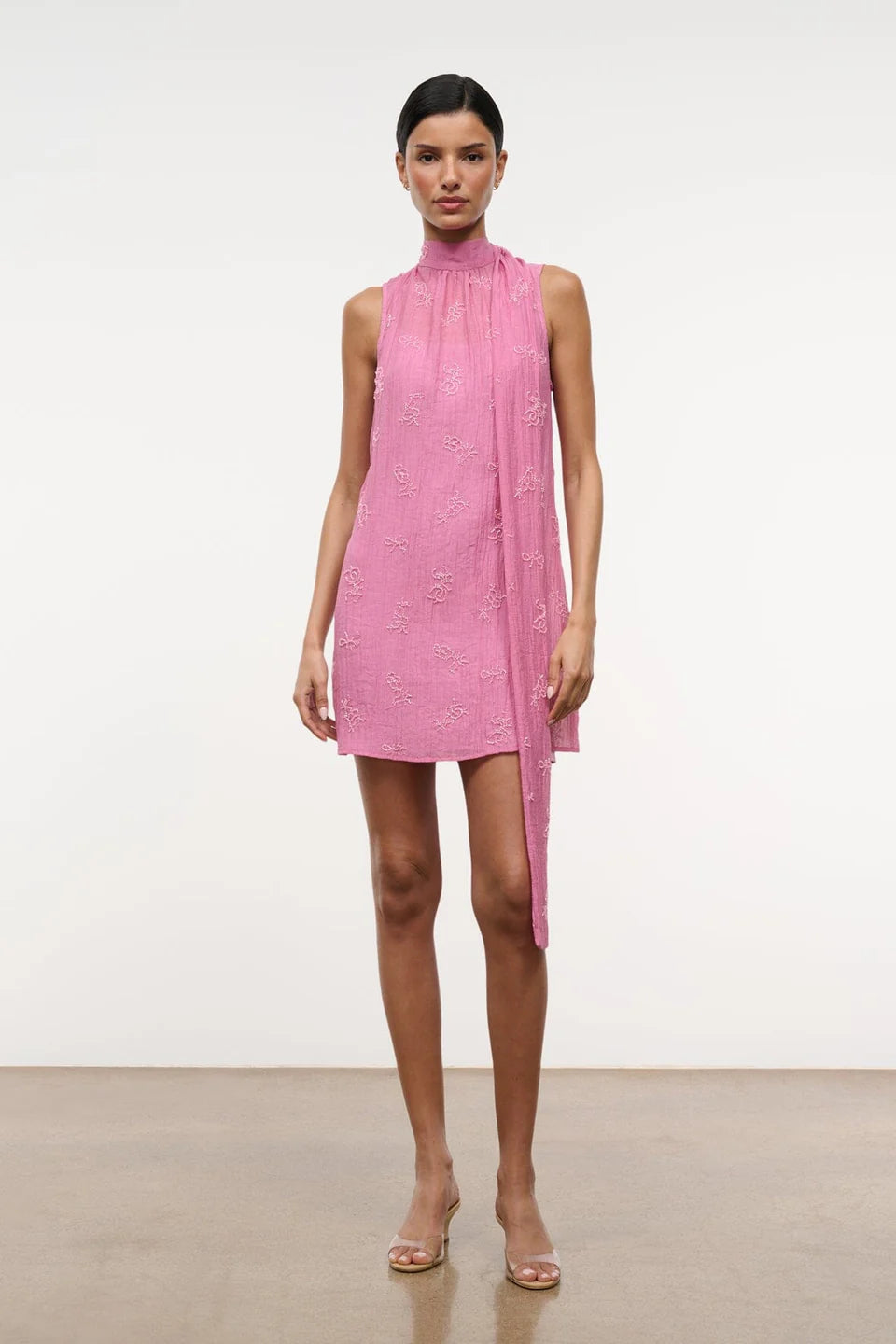 A model wears the Mini Albee Dress, an elegant pink sleeveless mini dress with a high mock neck and a draped fabric panel extending from the shoulder, creating a sophisticated silhouette.