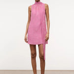 A model wears the Mini Albee Dress, an elegant pink sleeveless mini dress with a high mock neck and a draped fabric panel extending from the shoulder, creating a sophisticated silhouette.