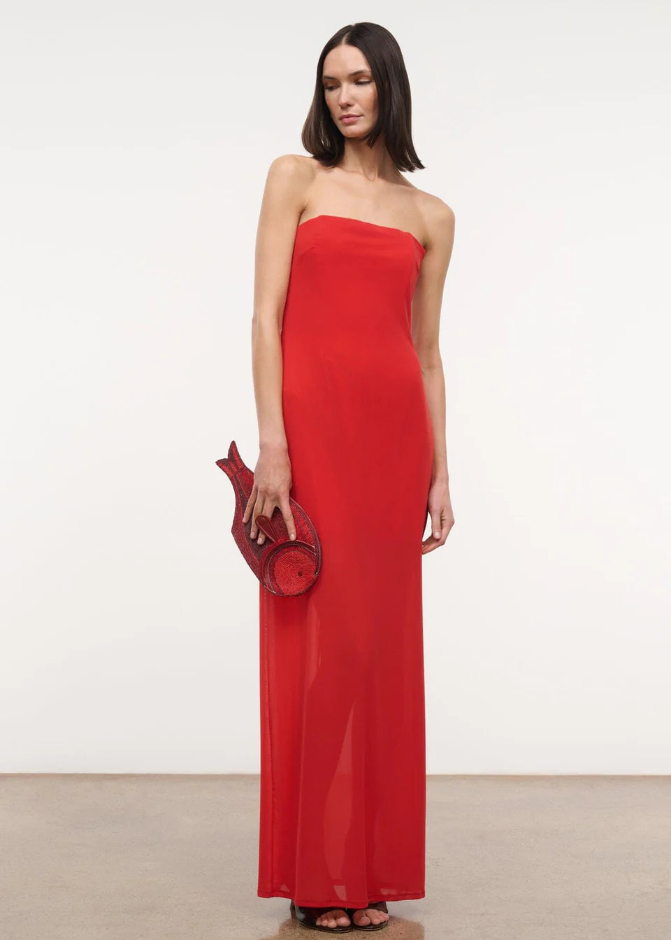 A person wears a red floor-length Liliana Dress styled with a matching fish-shaped clutch for a special occasion.