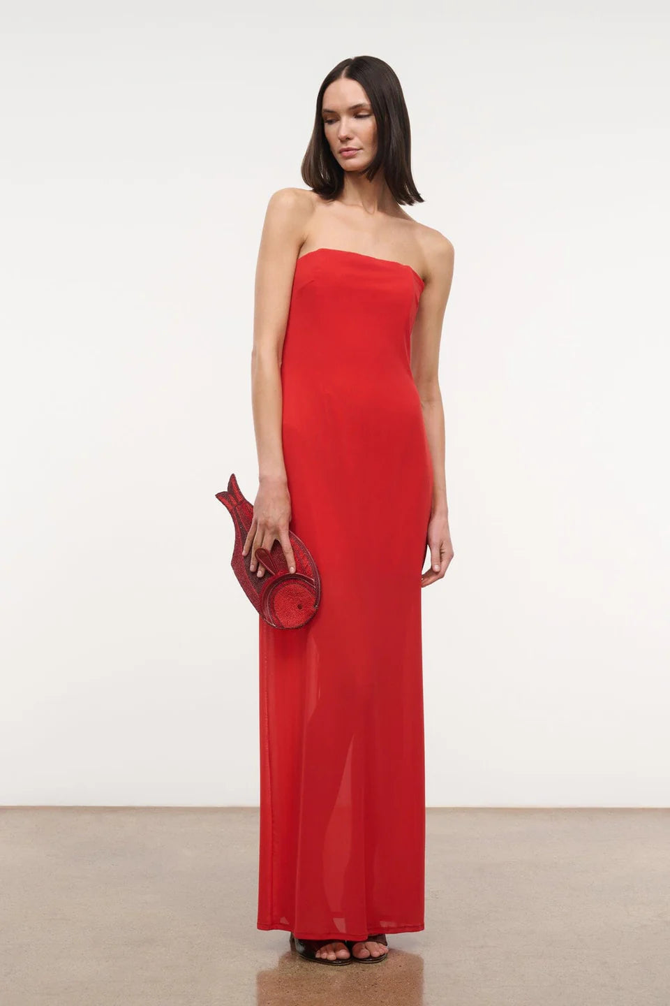 A person wears a red floor-length Liliana Dress styled with a matching fish-shaped clutch for a special occasion.