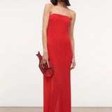 A person wears a red floor-length Liliana Dress styled with a matching fish-shaped clutch for a special occasion.