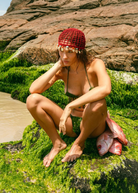 KABE SHOP INC Zahi Hat, a red crocheted skullcap adorned with seashells, worn by a model on a mossy coastal rock.