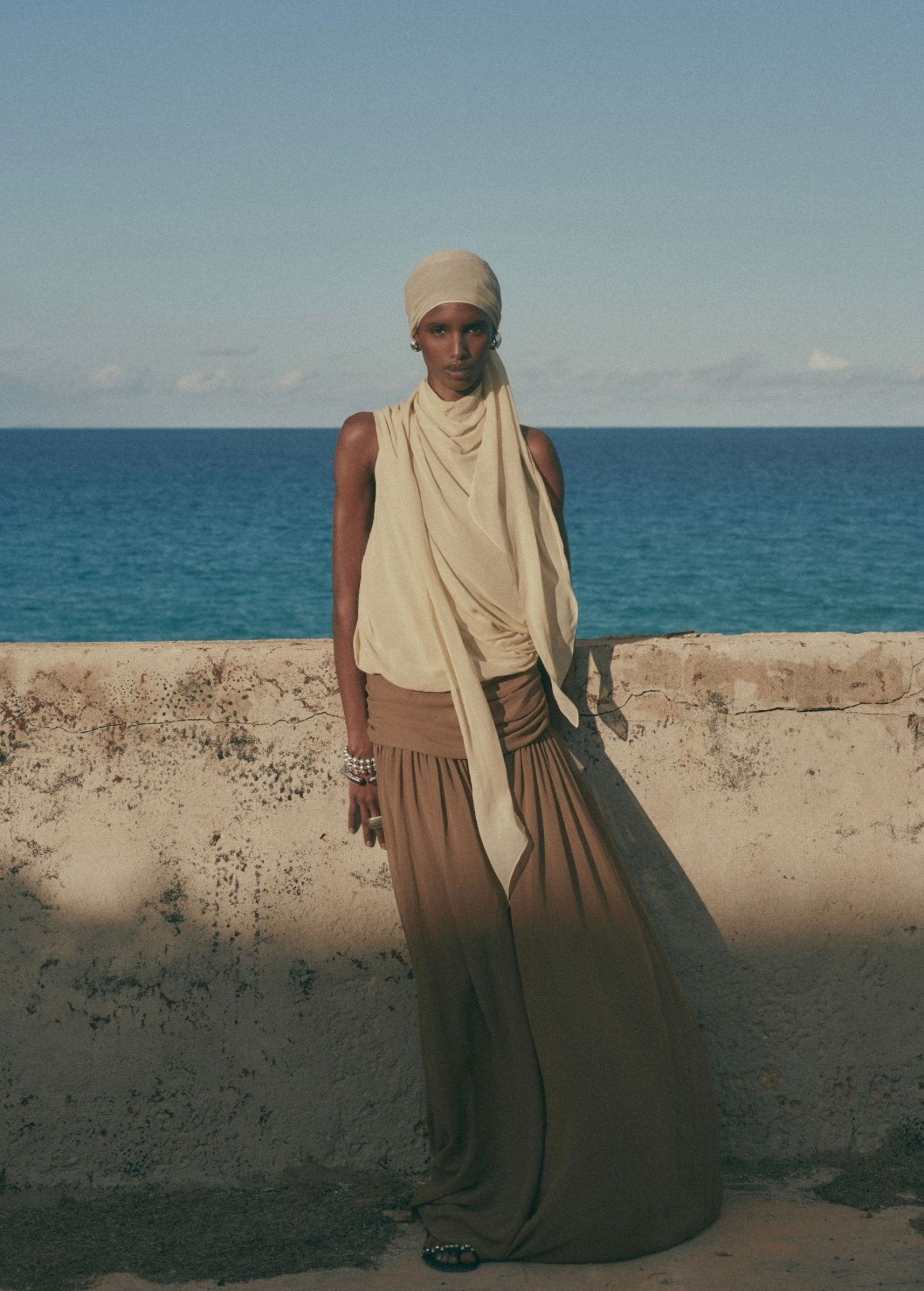 A person in a cream-colored headwrap and draped top paired with the Agapito Skirt in taupe chiffon, leaning against a stone wall with an ocean backdrop that creates an elongated silhouette.