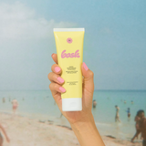 KABE SHOP INC SPF 50 sunscreen lotion in a yellow tube held by a hand against a blurred beach background.