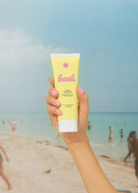 KABE SHOP INC SPF 50 sunscreen lotion in a yellow tube held by a hand against a blurred beach background.