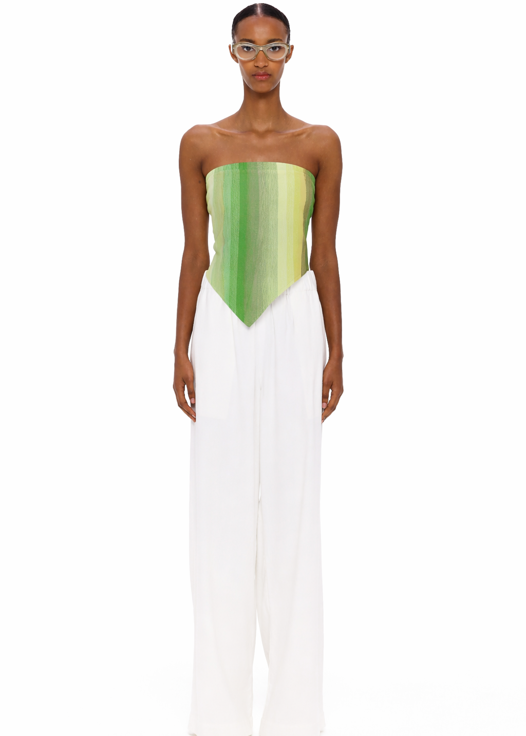 A person stands facing forward, wearing clear glasses, The Triangle Top—a green strapless top with a pointed hem crafted from handwoven textiles inspired by traditional Moroccan weaving—and loose white pants. The background is plain white.