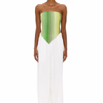 A person stands facing forward, wearing clear glasses, The Triangle Top—a green strapless top with a pointed hem crafted from handwoven textiles inspired by traditional Moroccan weaving—and loose white pants. The background is plain white.