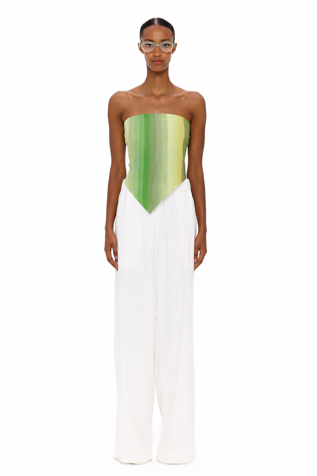 A person stands facing forward, wearing clear glasses, The Triangle Top—a green strapless top with a pointed hem crafted from handwoven textiles inspired by traditional Moroccan weaving—and loose white pants. The background is plain white.