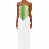 A person stands facing forward, wearing clear glasses, The Triangle Top—a green strapless top with a pointed hem crafted from handwoven textiles inspired by traditional Moroccan weaving—and loose white pants. The background is plain white.