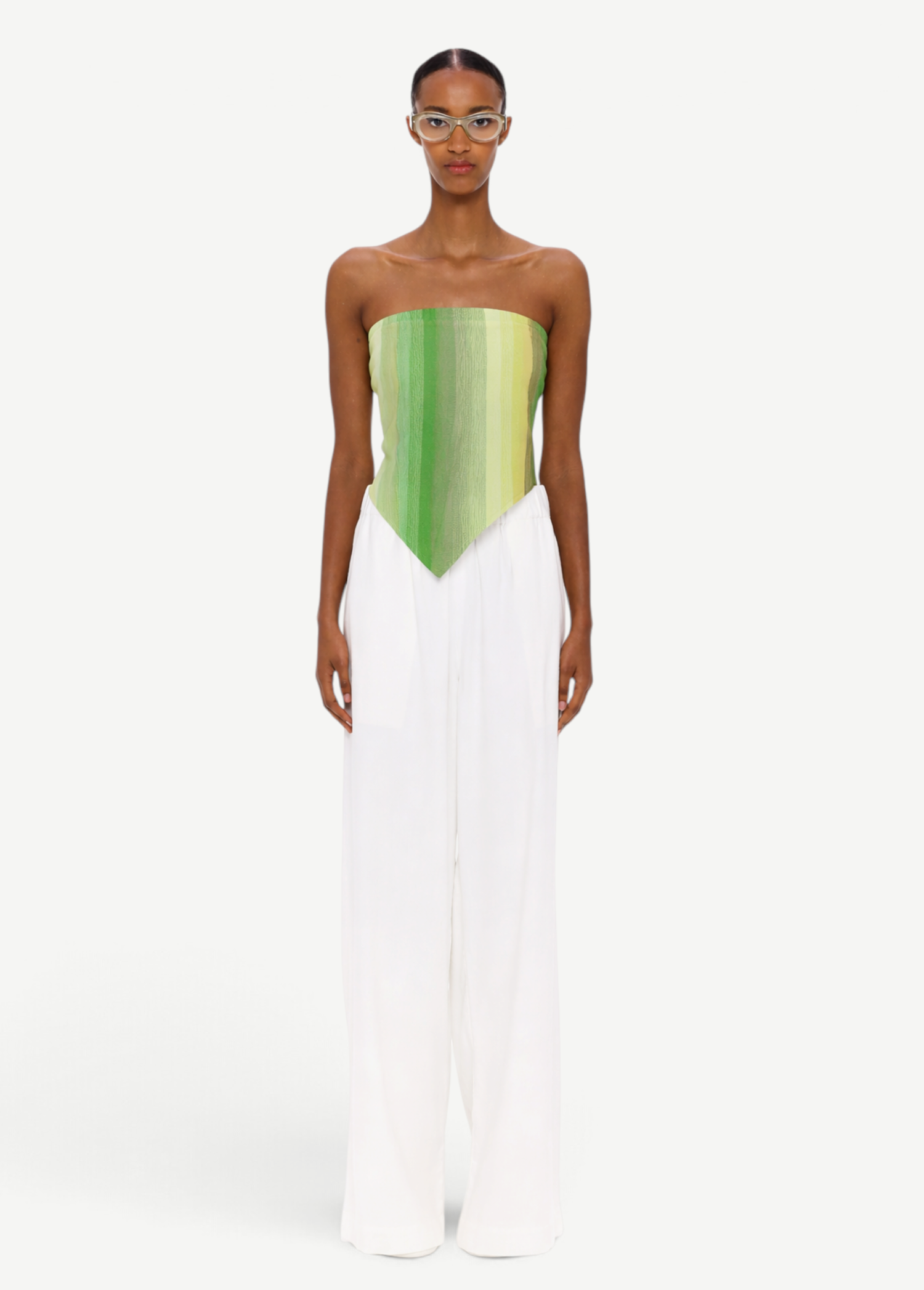A person stands facing forward, wearing clear glasses, The Triangle Top—a green strapless top with a pointed hem crafted from handwoven textiles inspired by traditional Moroccan weaving—and loose white pants. The background is plain white.