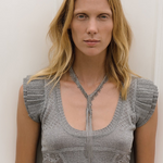 Wearing a short-sleeved, textured gray top, a person with long blonde hair stands against a plain white background looking directly at the camera, showcasing the Lariat Necklace White Bronze with its adjustable length.