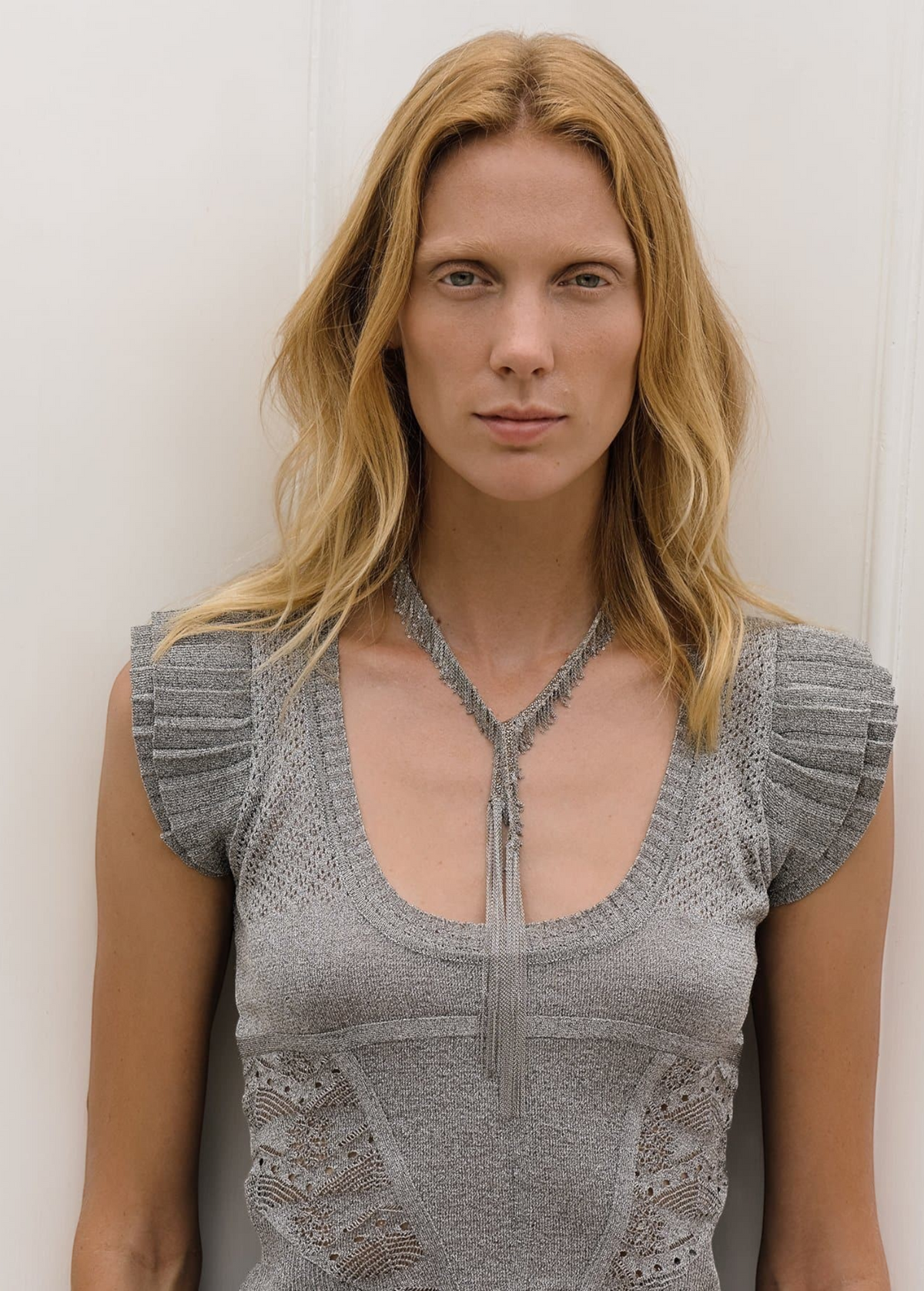 Wearing a short-sleeved, textured gray top, a person with long blonde hair stands against a plain white background looking directly at the camera, showcasing the Lariat Necklace White Bronze with its adjustable length.