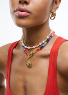 A woman wearing a red tank top and the Elsa Earrings—lightweight 14KT gold plate hoops—along with layered necklaces, one beaded with pearls and the other strung with colorful beads and a sun pendant, poses against a white background.