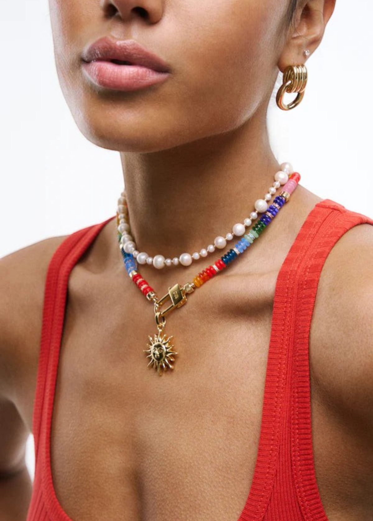 A woman wearing a red tank top and the Elsa Earrings—lightweight 14KT gold plate hoops—along with layered necklaces, one beaded with pearls and the other strung with colorful beads and a sun pendant, poses against a white background.