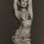 Wearing the Equator Top Swimwear, a woman poses with her hands behind her head, showcasing the leopard-print triangle bikini top made from recycled nylon and paired with loose jeans against a plain, dark background.