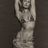 Wearing the Equator Top Swimwear, a woman poses with her hands behind her head, showcasing the leopard-print triangle bikini top made from recycled nylon and paired with loose jeans against a plain, dark background.
