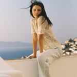 A woman in a handwoven crochet top and light-wash wide-leg Casa Pant sits on a white wall overlooking the sea.