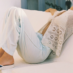 A person wearing wide-leg, light blue Casa Pant cropped trousers with lace detailing and a cream-colored, handwoven crochet cardigan, resting on a white surface.