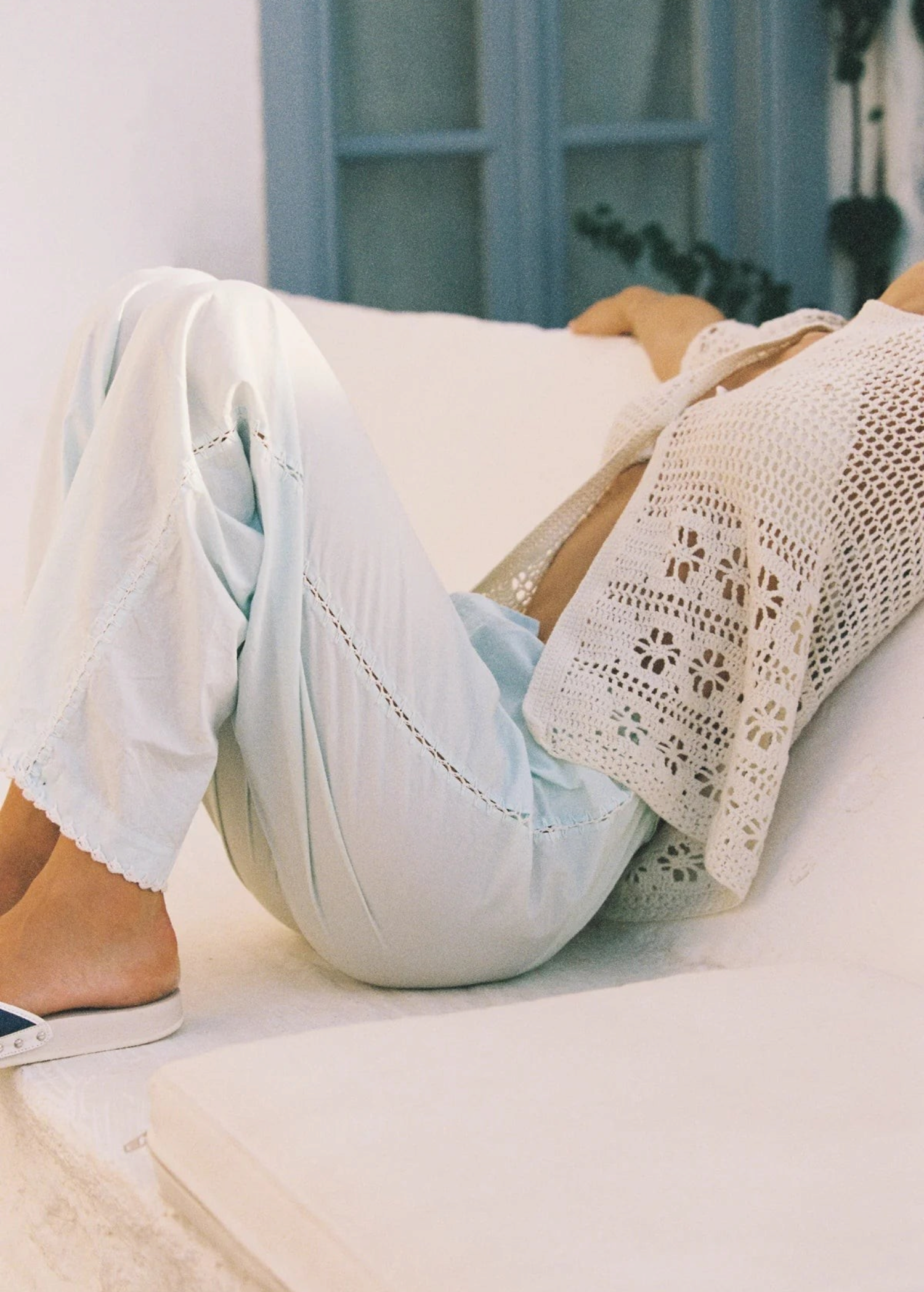 A person wearing wide-leg, light blue Casa Pant cropped trousers with lace detailing and a cream-colored, handwoven crochet cardigan, resting on a white surface.