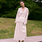 A person wearing an elegant Kiki Shirt with a long-sleeved, v-neck cut and a matching midi-length skirt, both featuring a vertical pinstripe pattern, standing outdoors on a wooden platform.