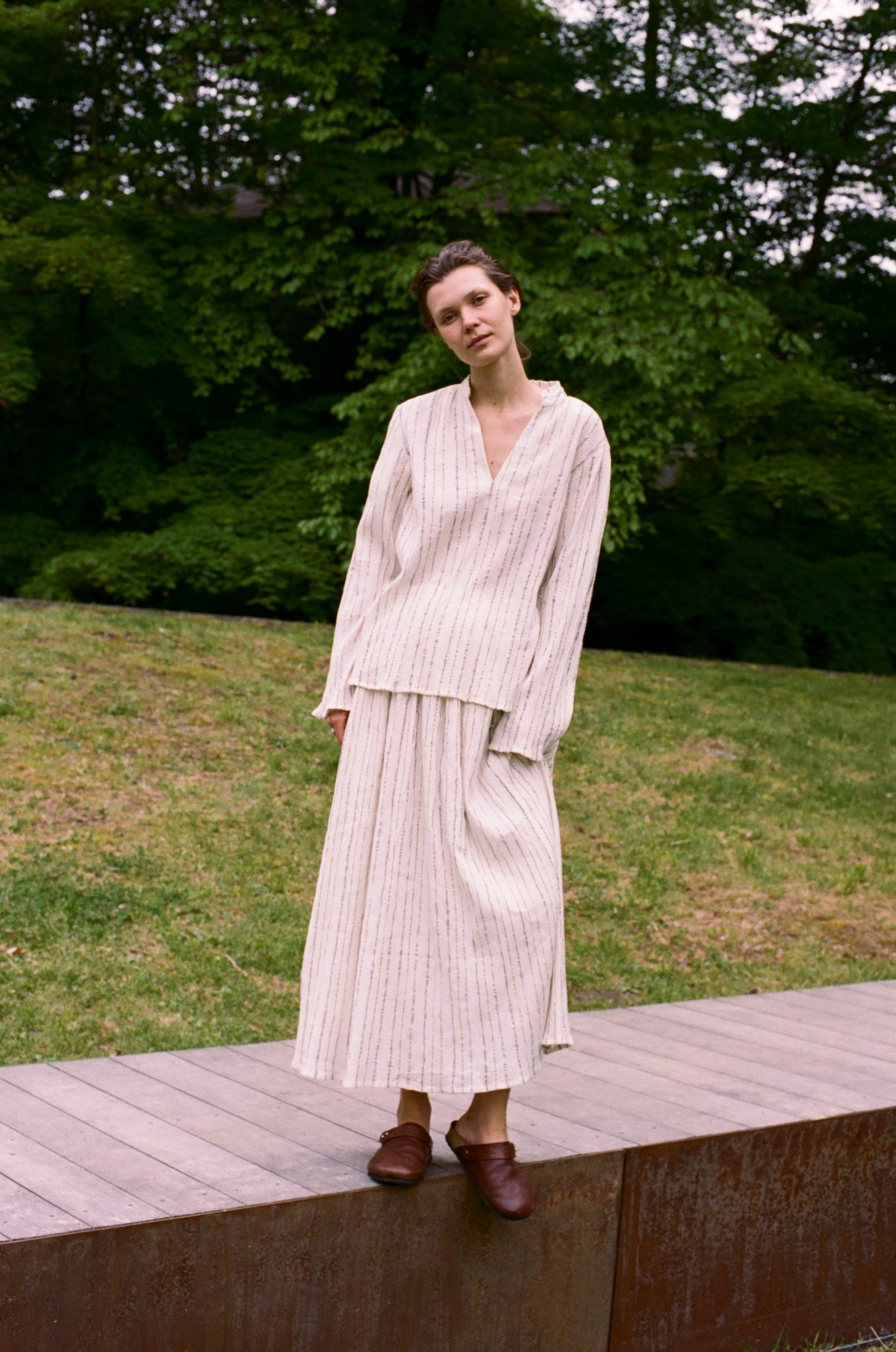 A person wearing an elegant Kiki Shirt with a long-sleeved, v-neck cut and a matching midi-length skirt, both featuring a vertical pinstripe pattern, standing outdoors on a wooden platform.