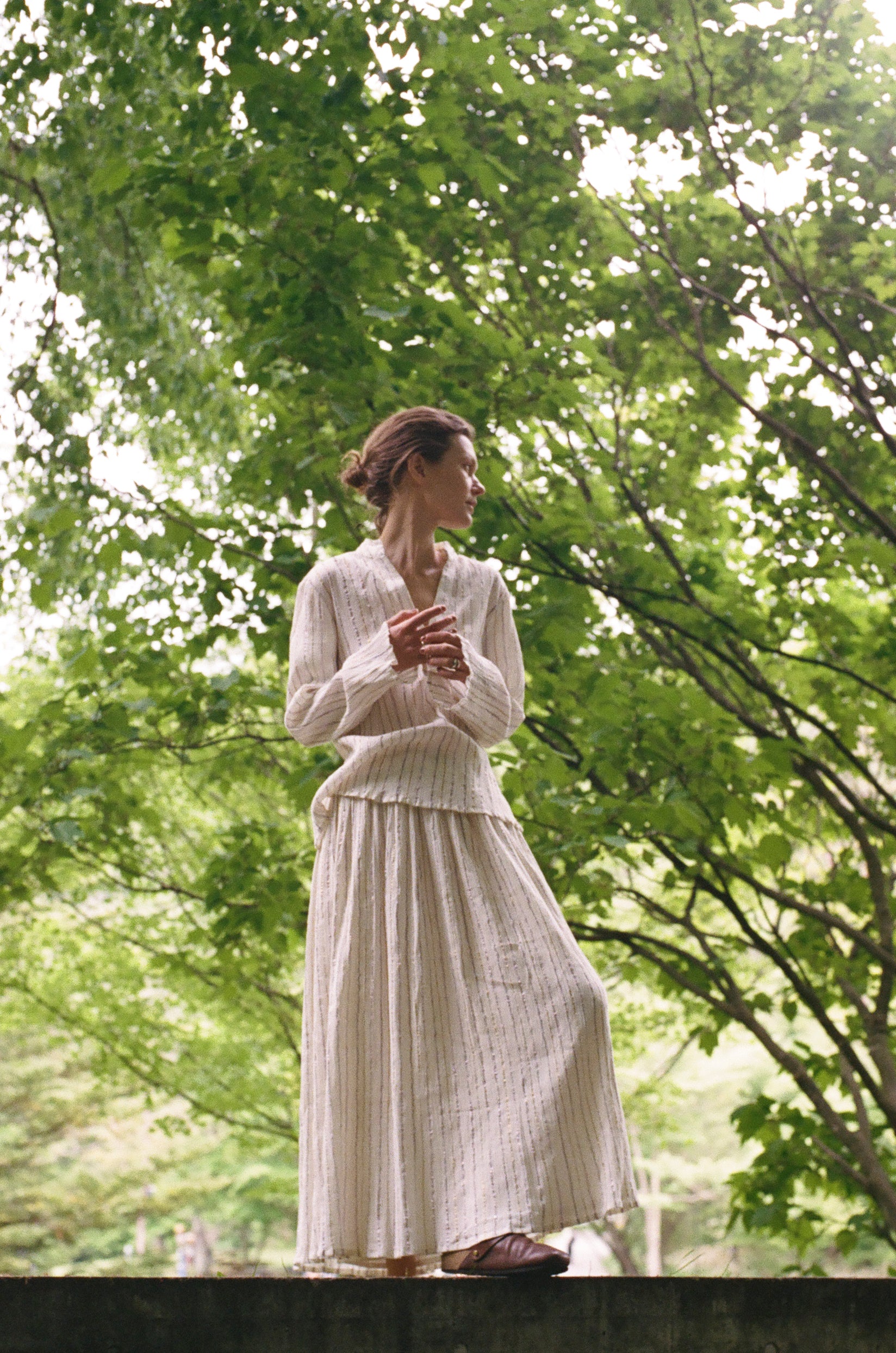 A person wears an elegant Kiki Shirt and a matching pleated midi skirt with thin vertical stripes, creating a romantic silhouette while standing outdoors among trees.