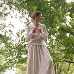 A woman stands in a wooded area wearing an elegant Kiki Shirt and a matching pleated maxi skirt with thin vertical stripes, creating a romantic silhouette.