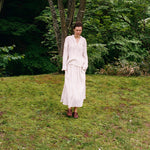 A person wearing an elegant pinstriped Kiki Shirt and matching long skirt stands on a grassy hill, creating a romantic silhouette.