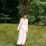A person wearing an elegant pinstriped Kiki Shirt with a matching long skirt stands on a grassy slope, creating a romantic silhouette.
