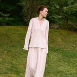 A woman wears an elegant cream Kiki Shirt with a v-neck and matching wide-leg trousers featuring thin vertical stripes, creating a romantic silhouette as she stands on a grassy slope.