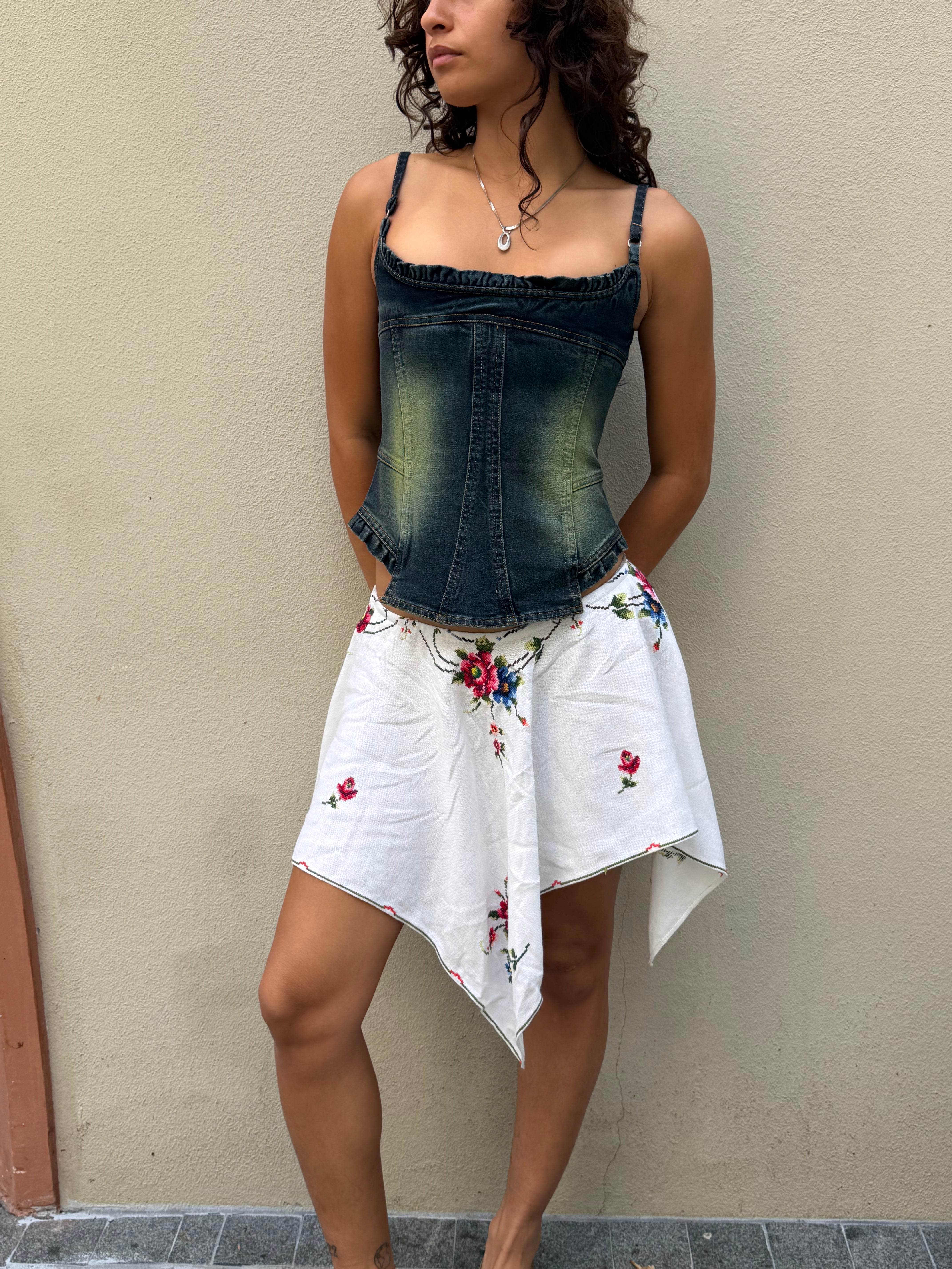 A woman stands against a beige wall wearing a denim corset top and the Celia Dragouni x KABE Embroidered Mini Skirt, her hands casually resting in her pockets.