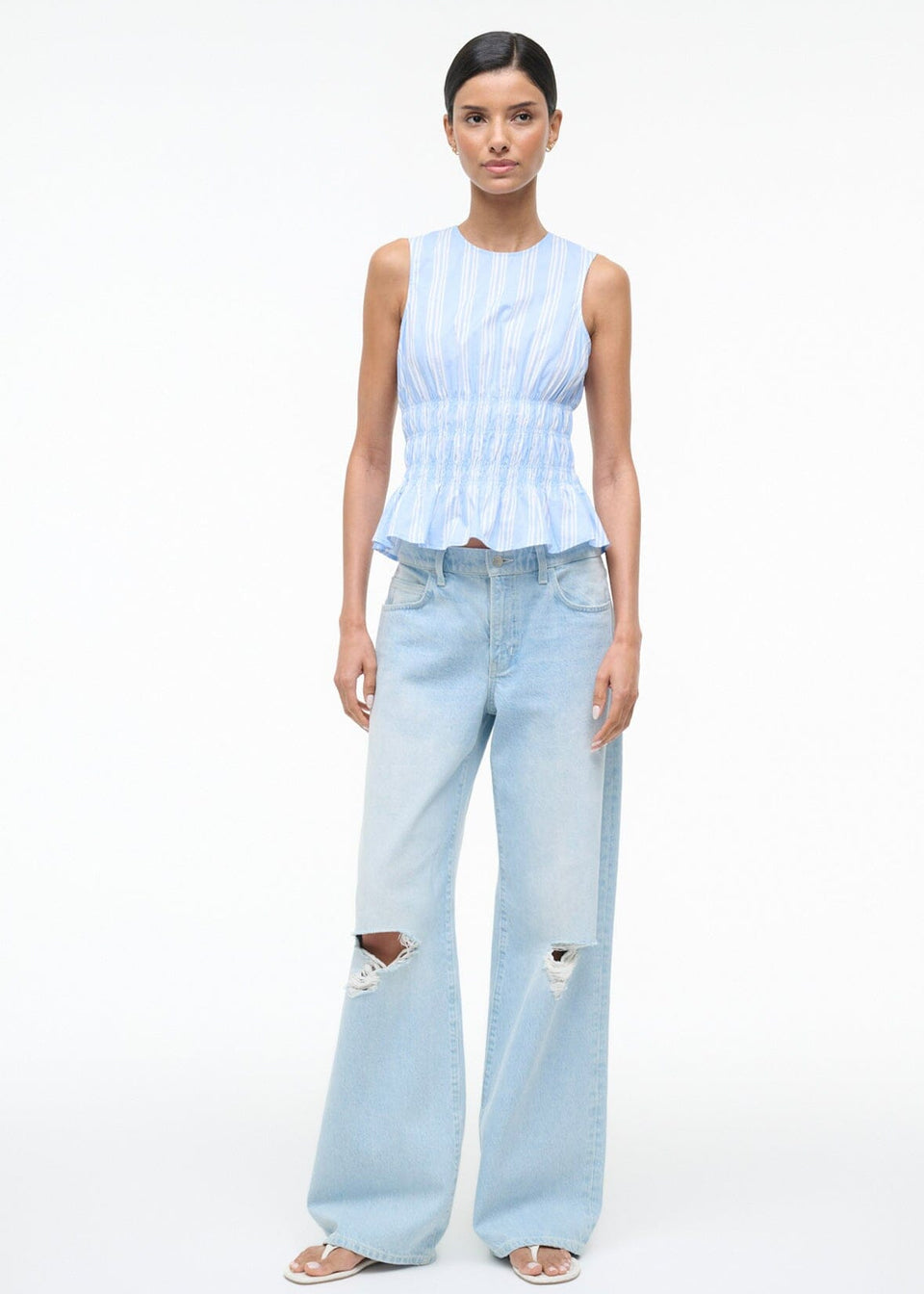 Culver Top featuring a sleeveless, blue-and-white striped peplum design with a smocked waist, paired with light-wash, ripped wide-leg jeans.