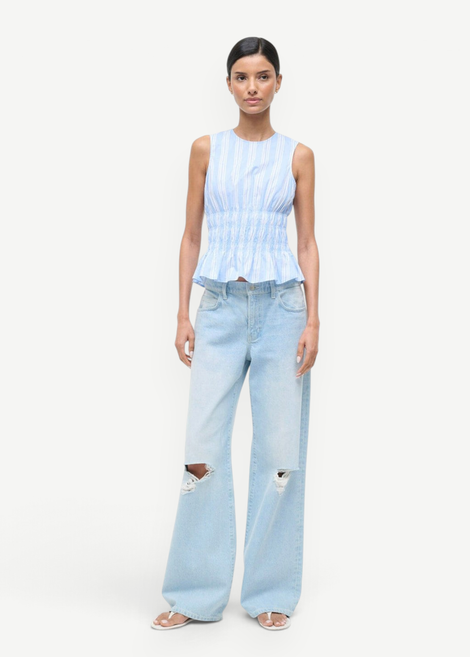 Culver Top featuring a sleeveless, blue-and-white striped peplum design with a smocked waist, paired with light-wash, ripped wide-leg jeans.