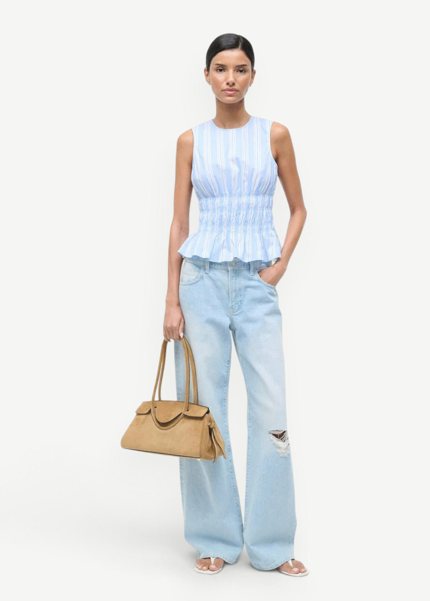 A model wears the light blue and white striped Culver Top with a sleeveless, smocked-waist silhouette, paired with light-wash distressed jeans and a tan handbag for a feminine style.