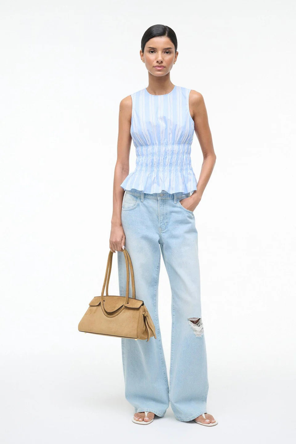 A model wears the light blue and white striped Culver Top with a sleeveless, smocked-waist silhouette, paired with light-wash distressed jeans and a tan handbag for a feminine style.
